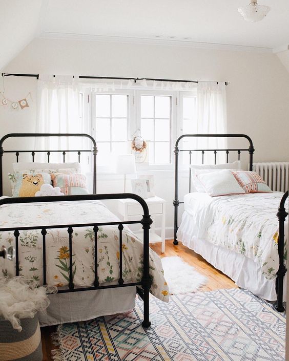 A light filled shared girls' bedroom with black metal beds with floral bedding, white curtains and a nightstand