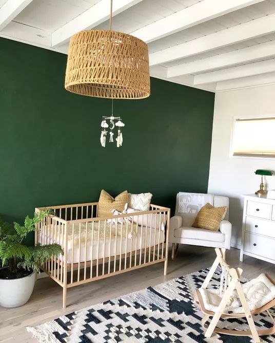 A mid century modern boho nursery with a green accent wall, white and light colored furniture, a printed rug and a woven lamp