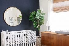 a mid-century modern nursery with navy walls, a white crib, a stained dresser, a round mirror, a bright printed rug and a gold chandelier