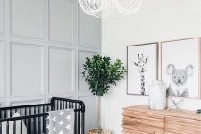a modern gender neutral nursery with a grey paneled wall, a wooden sideboard, a black crib and a catchy chandelier