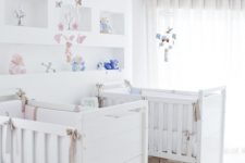 a neutral shared nursery with white furniture, open shelving, a crystal chandelier and pink and blue linens and toys