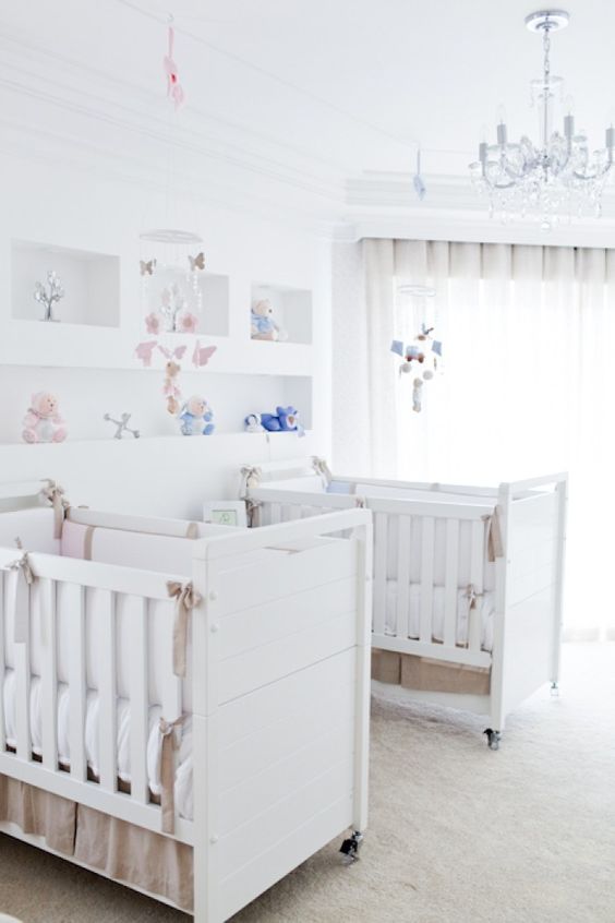 a neutral shared nursery with white furniture, open shelving, a crystal chandelier and pink and blue linens and toys