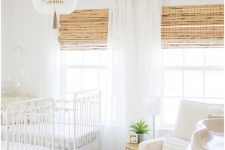 a neutral shared nursery with white furniture, woven shades, a beaded chandelier and a knit ottoman