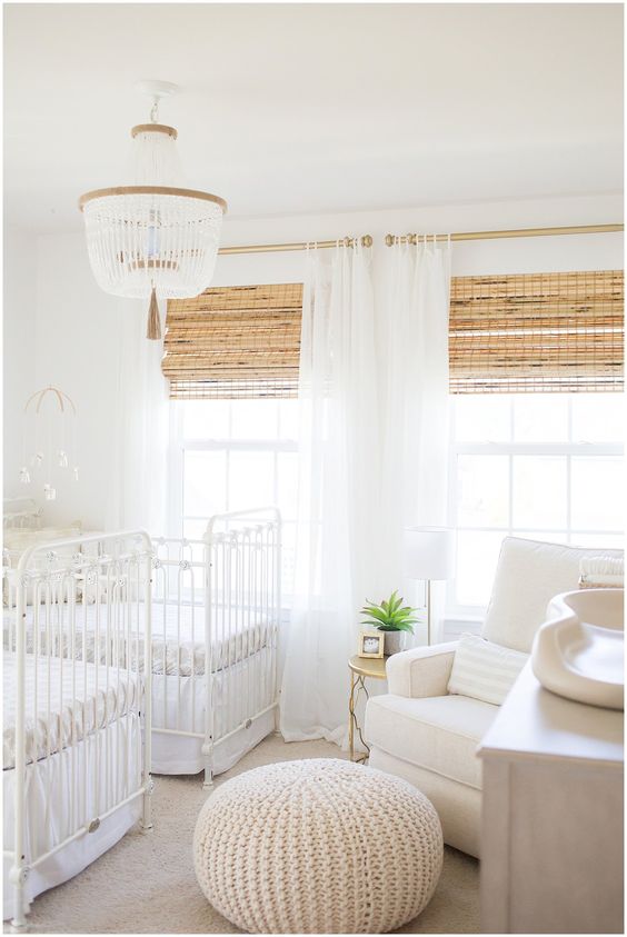 a neutral shared nursery with white furniture, woven shades, a beaded chandelier and a knit ottoman