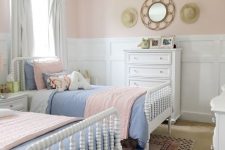 a pastel farmhouse shared girls’ bedroom with white paneling on the walls, white vintage beds, pink and blue bedding and a vintage chandelier
