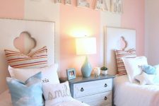 a pastel pink and white girls’ bedroom with a striped accent wall, marquee letters, white upholstered beds with pastel bedding, a blue and white dresser