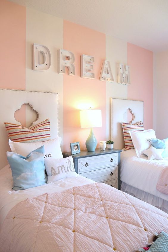 a pastel pink and white girls' bedroom with a striped accent wall, marquee letters, white upholstered beds with pastel bedding, a blue and white dresser