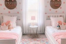 a pretty and bright shared girls’ bedroom with a polka dot accent wall, white farmhouse beds, pink and white bedding, a colorful rug and names