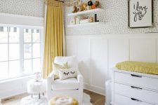 a pretty gender neutral nursery with a wallpaper and panel wall, white furniture, a woven lamp and touches of bright yellow