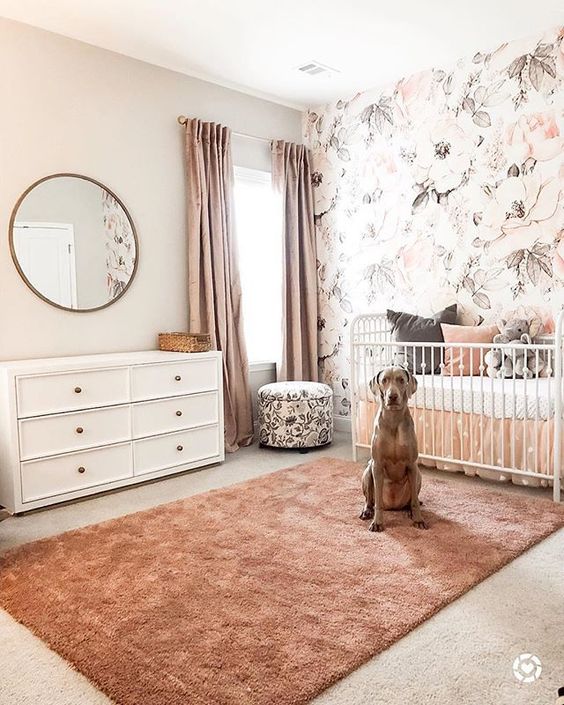 a pretty girlish nursery with a floral accent wall, white furniture, a floral ottoman, pink curtains and a peachy pink rug