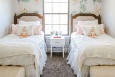 a pretty shared girls’ bedroom with a pink floral accent wall, pink upholstered beds, creamy poufs and a vintage chandelier