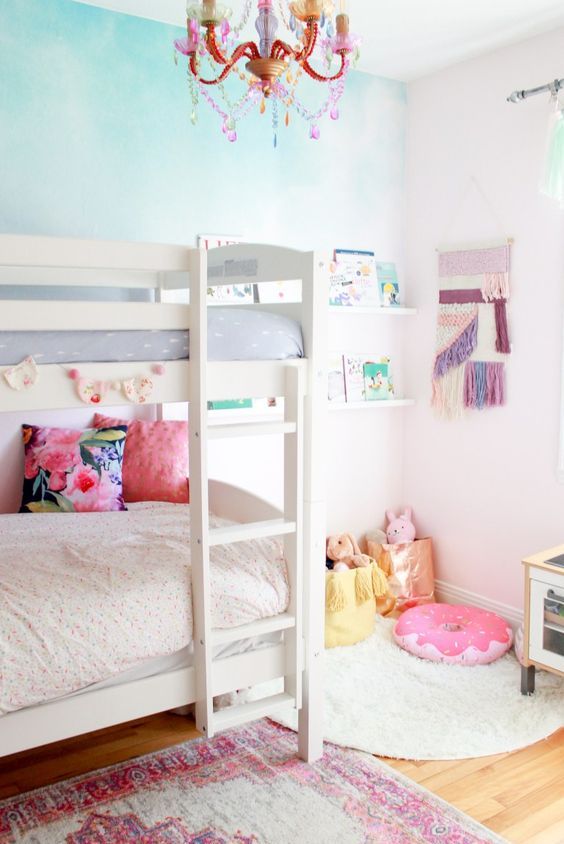 a pretty shared girls' bedroom with a white bunk bed, an ombre accent wall, bookshelves and a macrame hanging, toys and a bold rug