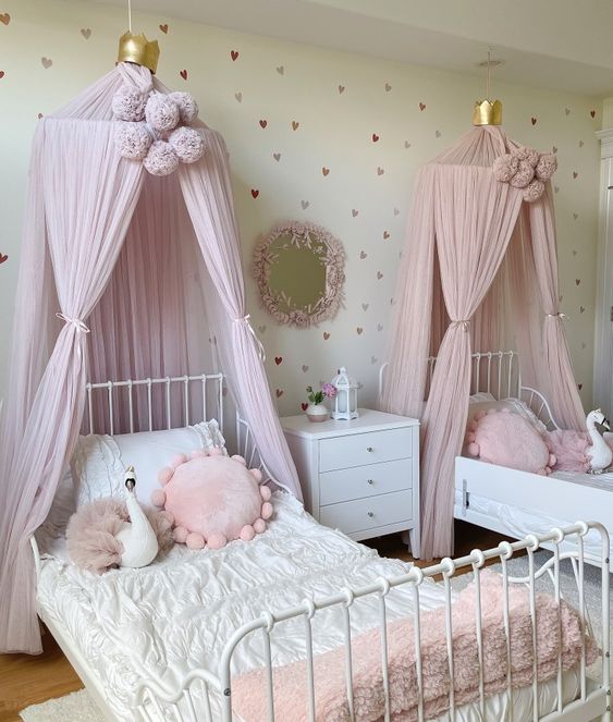 A princess style shared girls' bedroom with a white metal bed, pink canopies, pink pillows, a white nightstand and a mirror in a pink frame