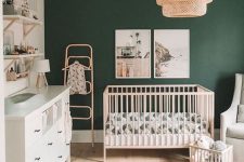 a stylish boho modern nursery with a hunter green accent wall, light-colored furniture, a woven chandelier and printed textiles