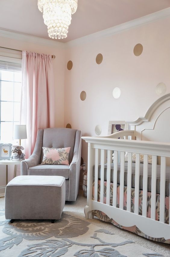 a stylish modern girl nursery with a polka dot wall, a white crib and a grey chair with an ottoman, a crystal chandelier and grey bedding