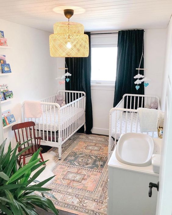 a stylish shared nursery with navy curtains, a boho rug, white furniture, a rattan lamp and cute cloud mobiles