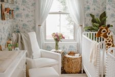 a tropical nursery with blue printed wallpaper, white furniture, a rattan stool, a beaded chandelier and a statement plant