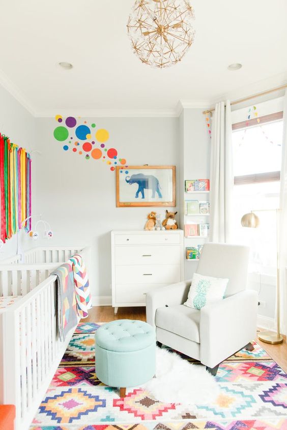 a vibrant shared nursery with white furniture, a colorful rug, ribbons and artworks on the wall and a floral chandelier