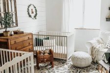 an elegant rustic nursery with white walls, a printed boho rug, white and stained furniture, a leather ottoman and a beaded chandelier