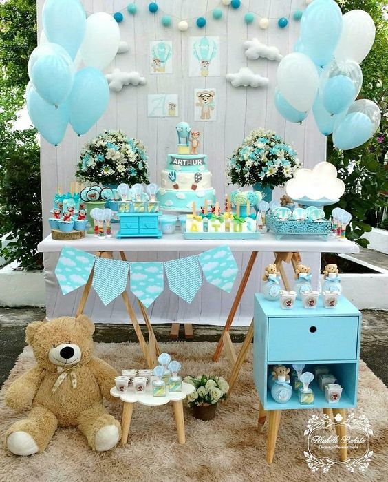 a blue dessert table with a backdrop done with clouds and blue balloons, a blue garland and blue and white floral arrangements
