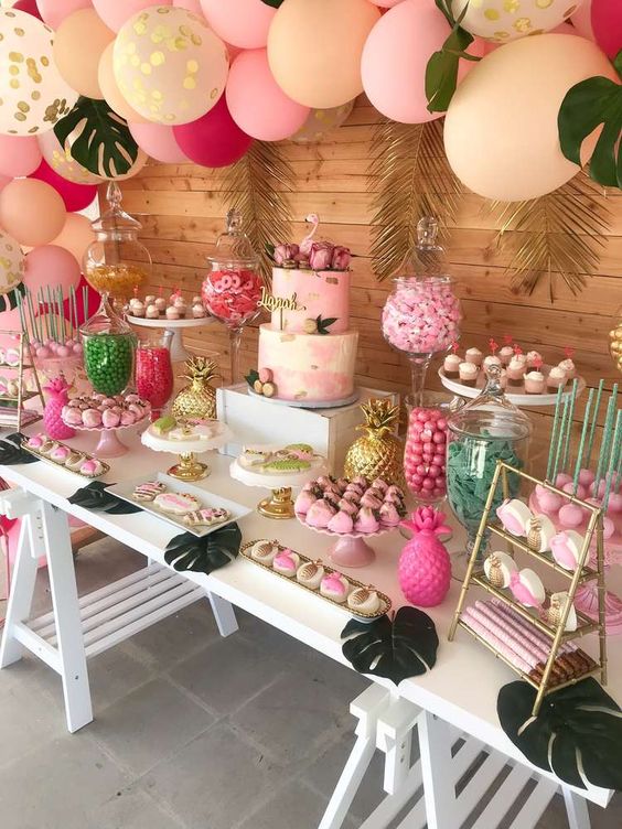 a bright pink, fold and fuchsia dessert table with a balloon garland over it, gilded tropical leaves, fake pineapples and jars and stands