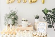 a neutral and gold baby shower dessert table with gold balloons, greenery arrangements and neutral desserts