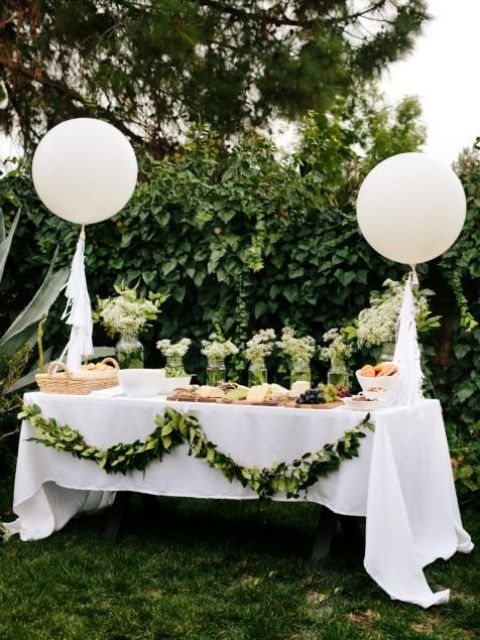 a neutral baby shower dessert table in white and green, with balloons and a greenery garland plus some neutral blooms