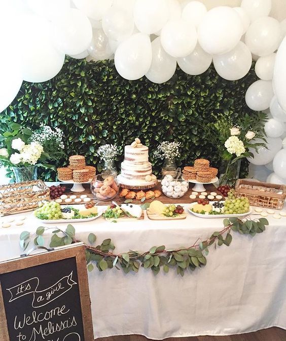 a neutral baby shower dessert table with a greenery backdrop, a large white balloon garland, a greenery garland and white blooms