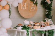 a neutral dessert table with a balloon garland with greenery and blooms, a name sign, a trestle table and greenery garlands