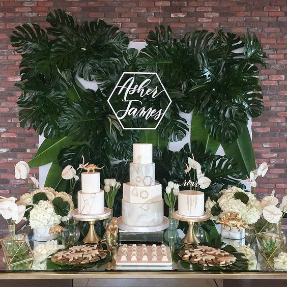 a neutral dessert table with a tropical feel, a lush tropical leaf wall, touches of gold, dinosaurs and white blooms