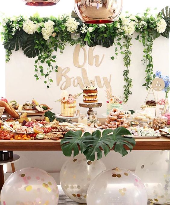 a neutral dessert table with lush greenery and white bloom garlands, large balloons with confetti, tropical leaves