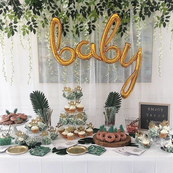 a tropical dessert table with gold calligraphy balloons, tropical greenery, a sign and lots of delicious sweets