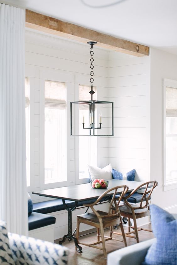 a coastal and rustic breakfast nook by the window with a dark table, rustic chairs, a pendant lamp and some blue and white pillows