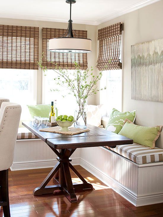 a farmhouse green and brown breakfast nook with a corner seating, a dark-stained table and various pillows, woven shades and green branches