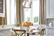 a cozy and elegant farmhouse breakfast nook with a built-in upholstered bench, a table and stained chairs, a pendant lamp and some blooms
