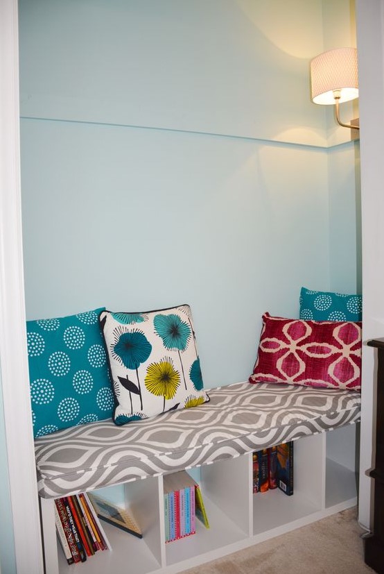 a Kallax piece turned into a sotrage bench for a reading nook, with pillows and colorful books, with a wall sconce