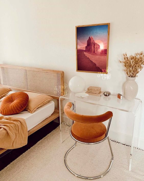 A catchy boho bedroom with a wod and cane bed, rust and peachy bedding, an acrylic desk, a rust colored curved chair and a bold artwork
