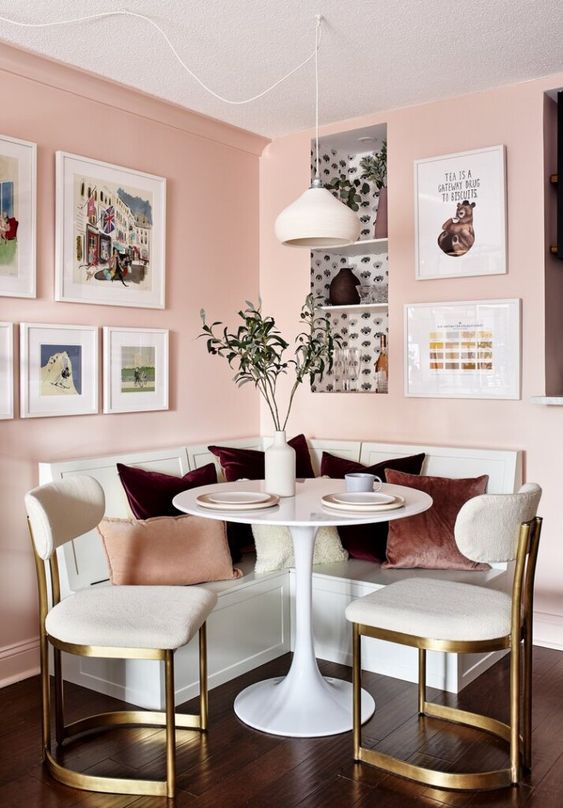 a chic modern breakfast nook with pink walls, gallery wall, a corner seating, white chairs, a table and a pendant lamp