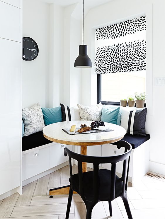 a contrasting breakfast corner with a corner seating and lots of pillows, a round table and a black chair, a blakc pendant lamp and a clock