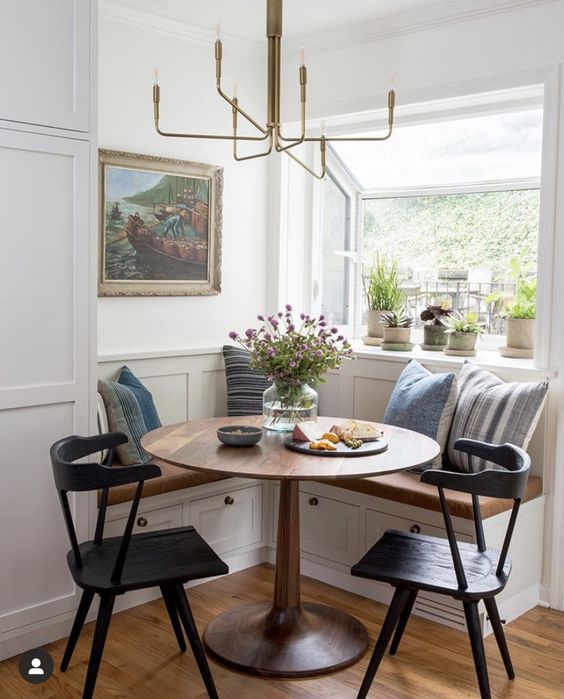 a cottage breakfast nook with a corner bench, a stained table and black chairs, a chandelier and potted plants plus an artwork