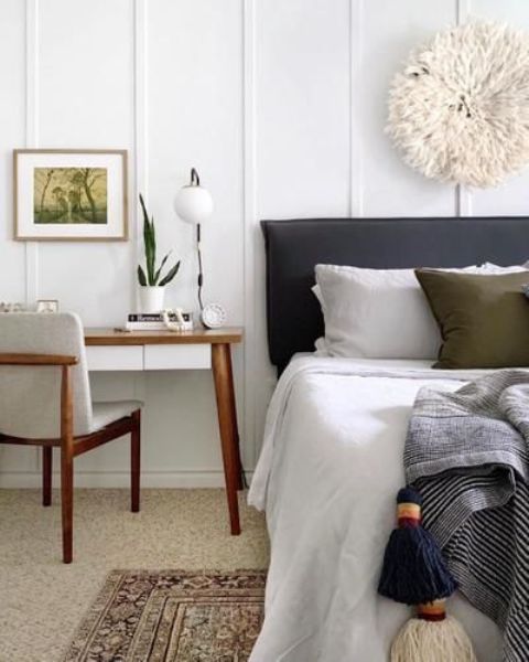 a farmhouse bedroom with a grey bed with neutral bedding, a white desk and a chair, some decor and layered rugs
