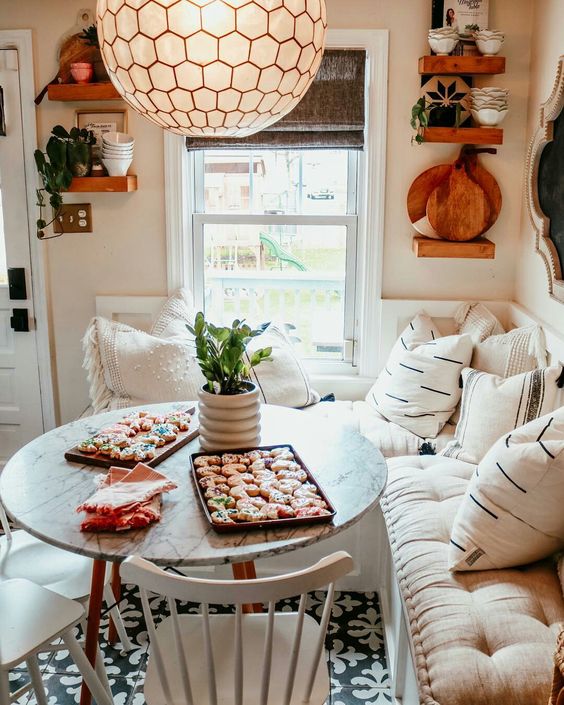 a modern farmohuse meets boho breakfast nook with a corner seat with pillows, a table and chairs, a pendant lamp and tableware and decor on the shelves