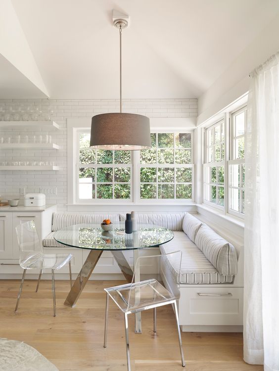 a neutral farmhouse breakfast corner with a large corner banquette seating, a glass table and acrylic chairs, a pendant lamp