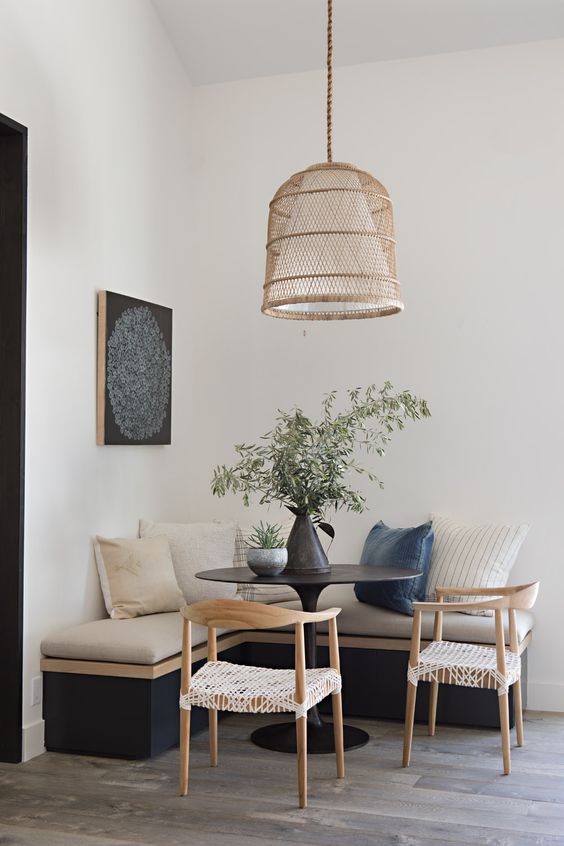 an earthy breakfast nook with a black corner seating and pillows, a black table and woven chairs, a pendant lamp and some greenery