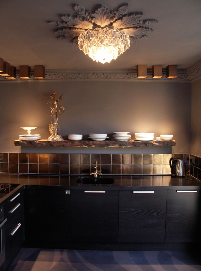The kitchen is modern, it's black cabinets and tiles contrast with a vintage chandelier