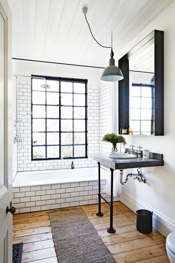 A retro industrial bathroom with the bathing zone clad with white subway tiles, a window with black frames, a mirror and a free standing sink