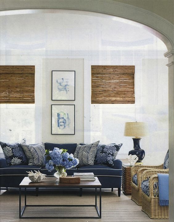 elegant beachy cottage living room design with soft sand walls paint color, bamboo roman shades, blue cushion sofa with white piping, brickmaker's coffee table, wicker chairs with blue cushions