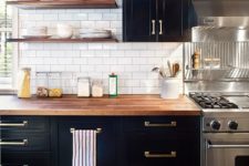 a classy black farmhouse kitchen with a white subway tile backsplash, butcherblock countertops and stained shelves plus brass handles is pure chic