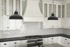 a classic white kitchen with a large statement hood, subway tile laid with a diagonal herringbone pattern, black countertops and pendant lamps