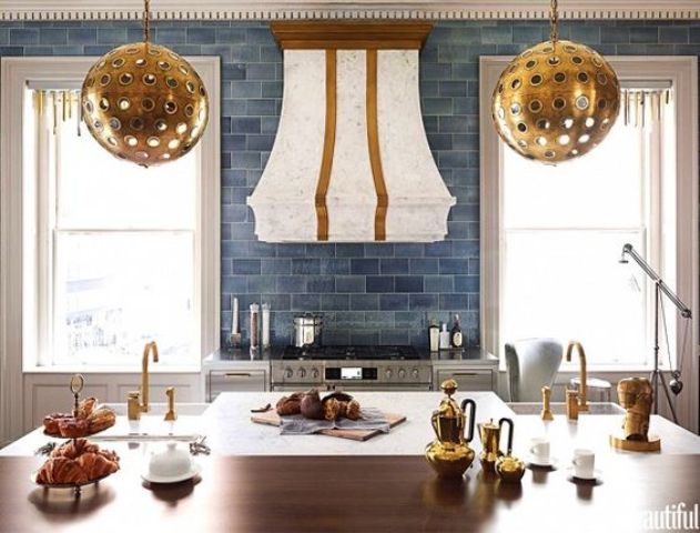 a glam and art deco kitchen with blue subway tiles of different shades, white and stained cabinets, a large hood and pendant lamps